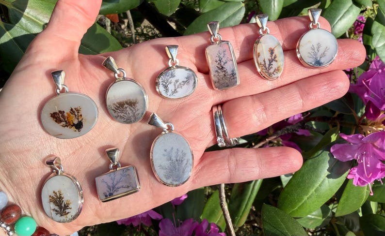 May include: A collection of silver pendants with dendritic agate stones. The pendants have various shapes, including oval, rectangular, and circular. The agate stones display unique, fern-like patterns in shades of brown, gray, and white. The pendants are displayed on an open hand.