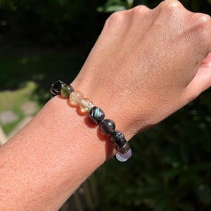May include: A beaded bracelet featuring a variety of natural gemstones in shades of black, green, yellow, and purple. The bracelet is strung on an elastic cord and is worn on a wrist.