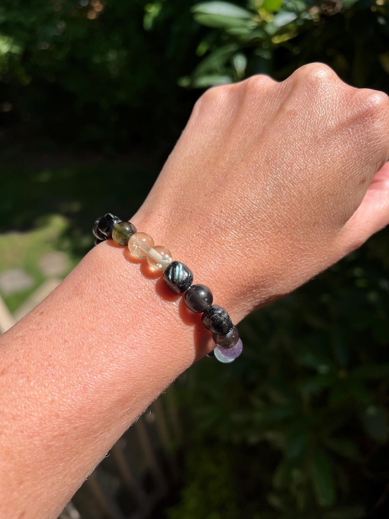 May include: A beaded bracelet featuring a variety of natural gemstones in shades of black, green, yellow, and purple. The bracelet is strung on an elastic cord and is worn on a wrist.
