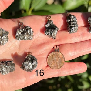 May include: Seven metallic, irregular, textured meteorite pendants with silver-toned bails, labeled with numbers 10, 14, 15, and 16. A copper-colored United States penny is included for scale, showing the relative size of these small, dark, jagged pieces.