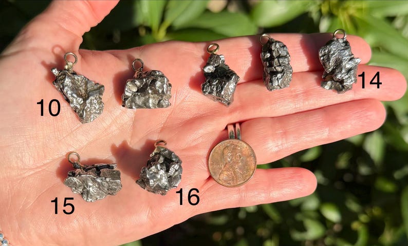 May include: Seven metallic, irregular, textured meteorite pendants with silver-toned bails, labeled with numbers 10, 14, 15, and 16. A copper-colored United States penny is included for scale, showing the relative size of these small, dark, jagged pieces.
