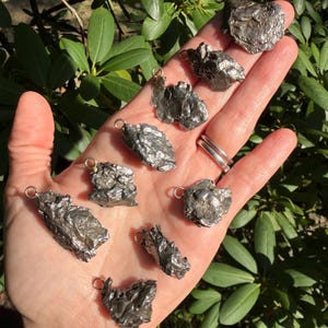 May include: A collection of eight irregular, metallic-toned meteorite pendants, each featuring a small circular gold-colored bail attached to the top. The surfaces of these pendants are textured, pitted, and display a dark, reflective gray finish.
