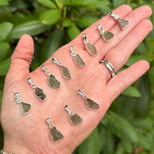 May include: A collection of ten small, raw, olive-green moldavite pendants, each set in a simple silver-toned metal bail. The stones feature irregular, textured surfaces and are arranged in a scattered pattern across the palm of a hand.