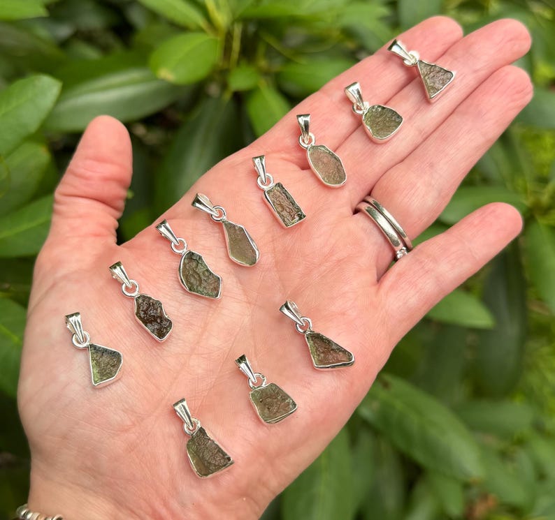 May include: A collection of ten small, raw, olive-green moldavite pendants, each set in a simple silver-toned metal bail. The stones feature irregular, textured surfaces and are arranged in a scattered pattern across the palm of a hand.