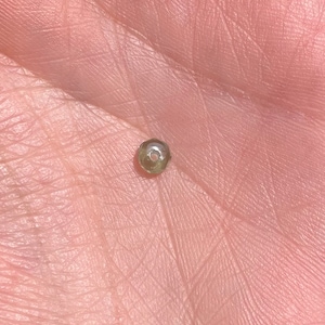 May include: A small, faceted, olive-green bead. The bead is held in a person's palm, showcasing its intricate cut and subtle color. The bead's surface reflects light, highlighting its multiple facets. This bead is suitable for jewelry making.