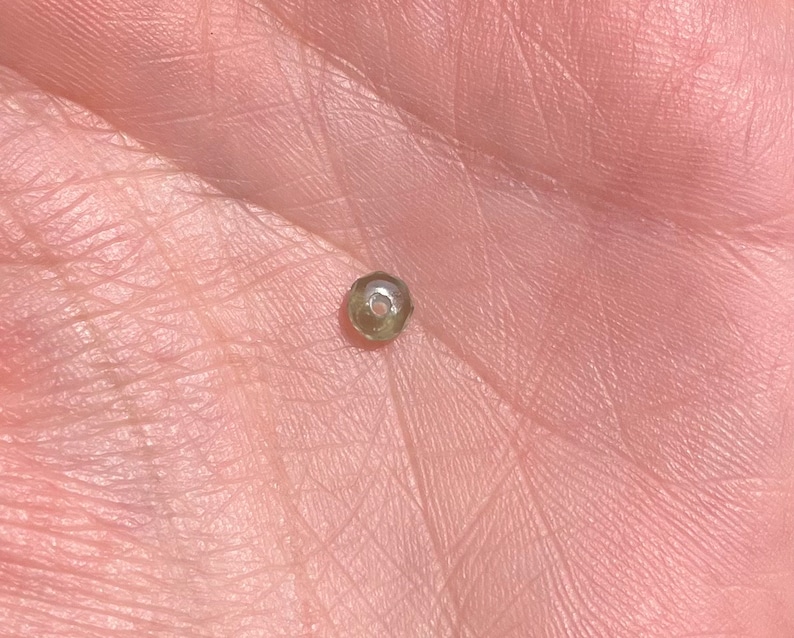 May include: A small, faceted, olive-green bead. The bead is held in a person's palm, showcasing its intricate cut and subtle color. The bead's surface reflects light, highlighting its multiple facets. This bead is suitable for jewelry making.