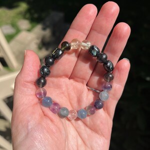 May include: A beaded bracelet featuring a variety of colorful gemstones. The bracelet includes black, yellow, green, purple, and blue beads. The bracelet is held in a hand, with the background out of focus.