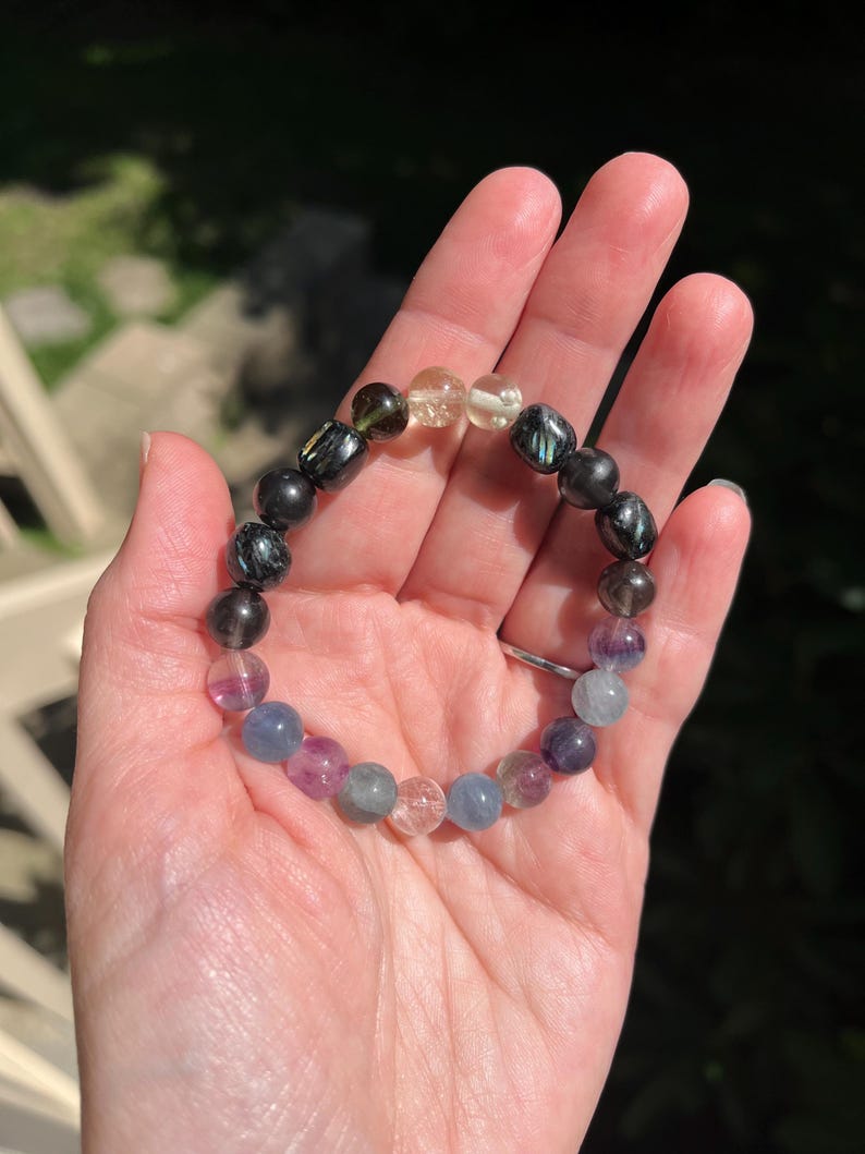 May include: A beaded bracelet featuring a variety of colorful gemstones. The bracelet includes black, yellow, green, purple, and blue beads. The bracelet is held in a hand, with the background out of focus.