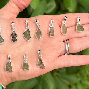 May include: A collection of eleven small, raw, olive-green moldavite pendants, each set in a simple silver-toned metal bail. The pendants vary in their irregular, textured shapes and shades of green, displayed together in a palm.