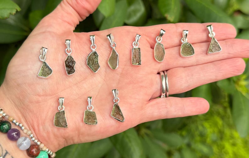 May include: A collection of eleven small, raw, olive-green moldavite pendants, each set in a simple silver-toned metal bail. The pendants vary in their irregular, textured shapes and shades of green, displayed together in a palm.