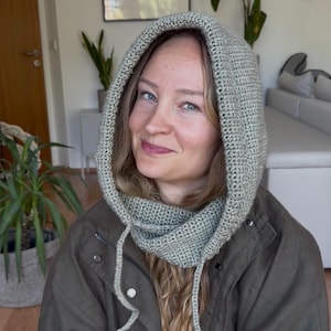 May include: A sage green crochet hood with a textured knit pattern and long matching ties. The hood is designed to cover the head and neck, featuring a thick, woven construction that frames the face with a soft, uniform stitch throughout.
