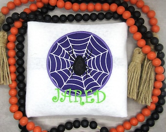 Puede incluir: Una almohada blanca con una telaraña morada y una araña negra en el centro. La almohada está rodeada por una guirnalda de cuentas negras y naranjas con borlas. El nombre "JARED" está bordado en verde debajo de la telaraña.