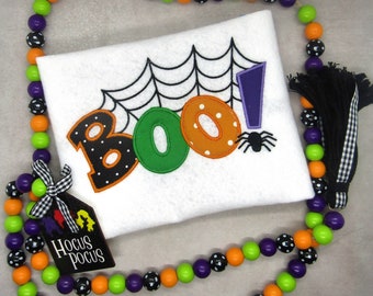 Puede incluir: Una tela blanca con la palabra "BOO!" bordada en letras negras, verdes y naranjas con lunares blancos. Una araña negra con un signo de exclamación morado está encima de la palabra. Una guirnalda de cuentas negras y blancas con cuentas naranjas rodea la tela. Un letrero de madera con las palabras "Hocus Pocus" está en la esquina inferior izquierda.