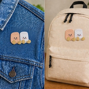 May include: Embroidered patch showing two cartoon marshmallows with smiling faces and gold-coloured feet. The patch is displayed on a denim jacket and a beige backpack, suggesting its use as an accessory for clothing or bags.