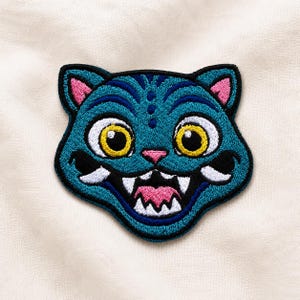 May include: An embroidered patch featuring the head of a teal cat with large yellow eyes, pink inner ears, and a pink nose. The cat has a wide, open mouth showing white teeth and a pink tongue, outlined in black thread on a textured fabric.