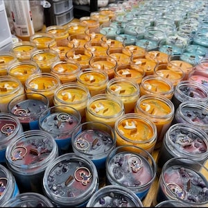 Puede incluir: Una colecci&oacute;n de velas perfumadas en frascos de vidrio transparente. Las velas presentan varios colores, incluyendo naranja, azul y morado, con elementos decorativos como cristales y amuletos. Algunas velas tienen mechas de madera.