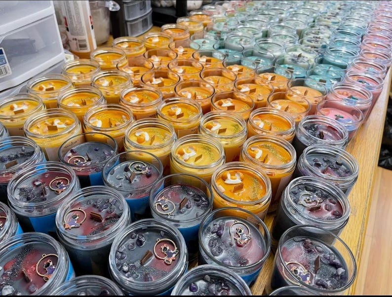 Puede incluir: Una colecci&oacute;n de velas perfumadas en frascos de vidrio transparente. Las velas presentan varios colores, incluyendo naranja, azul y morado, con elementos decorativos como cristales y amuletos. Algunas velas tienen mechas de madera.