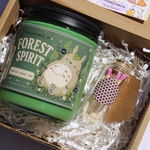 May include: A green candle jar with the text "FOREST SPIRIT" and a cartoon character illustration, inside a cardboard box with paper filler. A small glass jar of matches with pink tips is also in the box.