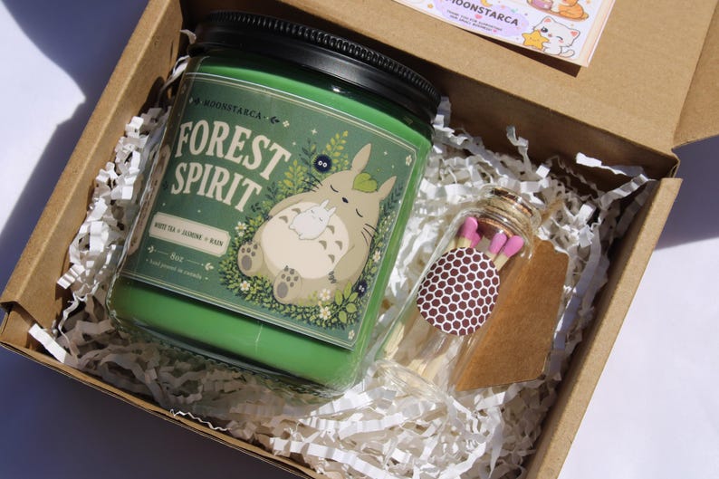 May include: A green candle jar with the text "FOREST SPIRIT" and a cartoon character illustration, inside a cardboard box with paper filler. A small glass jar of matches with pink tips is also in the box.