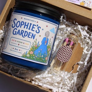 May include: A blue candle in a jar with a black lid, labeled "Sophie's Garden" with a floral illustration, and a small glass bottle of matches, all inside a cardboard box with paper filler.