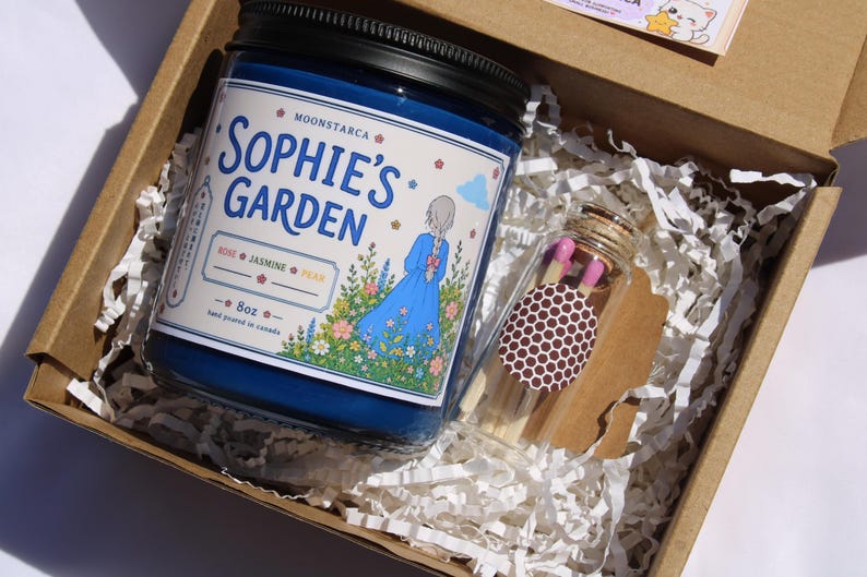 May include: A blue candle in a jar with a black lid, labeled "Sophie's Garden" with a floral illustration, and a small glass bottle of matches, all inside a cardboard box with paper filler.