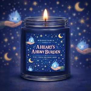 May include: A lit, navy blue jar candle with the text "A Heart's A Heavy Burden." The label features a starry night design with crescent moons and small blue spirit-like figures. The candle is 8 oz and scented with white tea, rose, and musk.