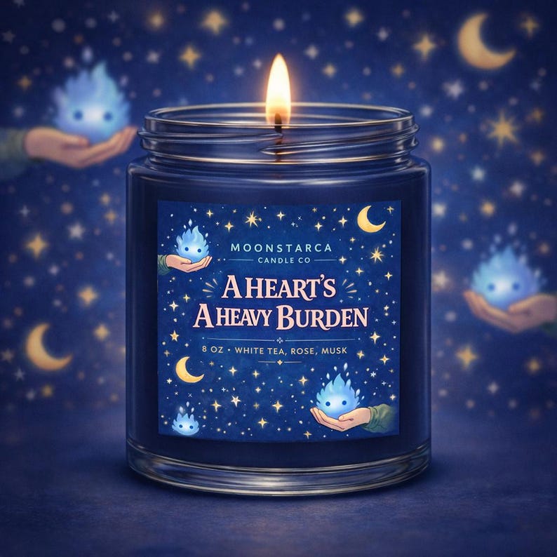 May include: A lit, navy blue jar candle with the text "A Heart's A Heavy Burden." The label features a starry night design with crescent moons and small blue spirit-like figures. The candle is 8 oz and scented with white tea, rose, and musk.