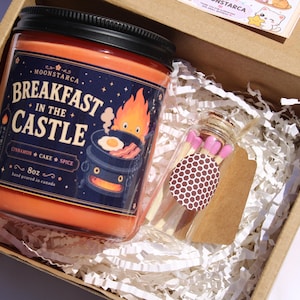 May include: A gift box containing a jar candle and a small glass bottle of matches. The candle is orange with a black lid and a label that reads "Breakfast in the Castle." The match bottle has a brown and white polka dot label.
