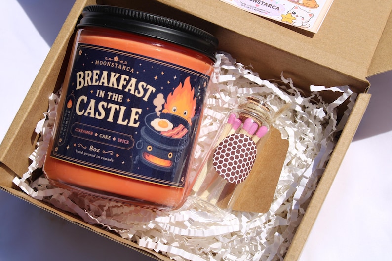 May include: A gift box containing a jar candle and a small glass bottle of matches. The candle is orange with a black lid and a label that reads "Breakfast in the Castle." The match bottle has a brown and white polka dot label.