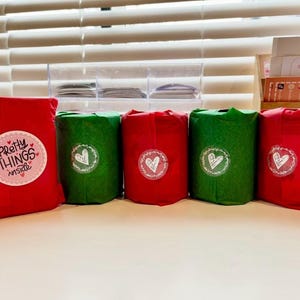 Puede incluir: Una colecci&oacute;n de regalos envueltos en rojo y verde, dispuestos sobre una superficie blanca. Los regalos est&aacute;n adornados con pegatinas con corazones y el texto "Pretty Things Inside". El fondo incluye una persiana y contenedores de almacenamiento.