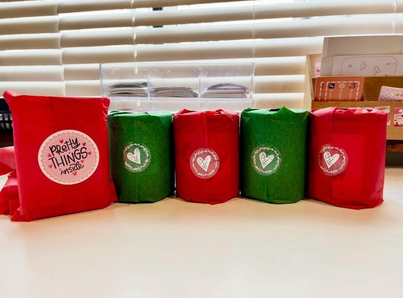 Puede incluir: Una colecci&oacute;n de regalos envueltos en rojo y verde, dispuestos sobre una superficie blanca. Los regalos est&aacute;n adornados con pegatinas con corazones y el texto "Pretty Things Inside". El fondo incluye una persiana y contenedores de almacenamiento.