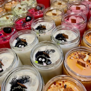 May include: A collection of various scented candles in clear glass jars. The candles feature a range of colors, including pink, red, white, and orange. Some candles have decorative elements like crystals, wood wicks, and cartoon characters.