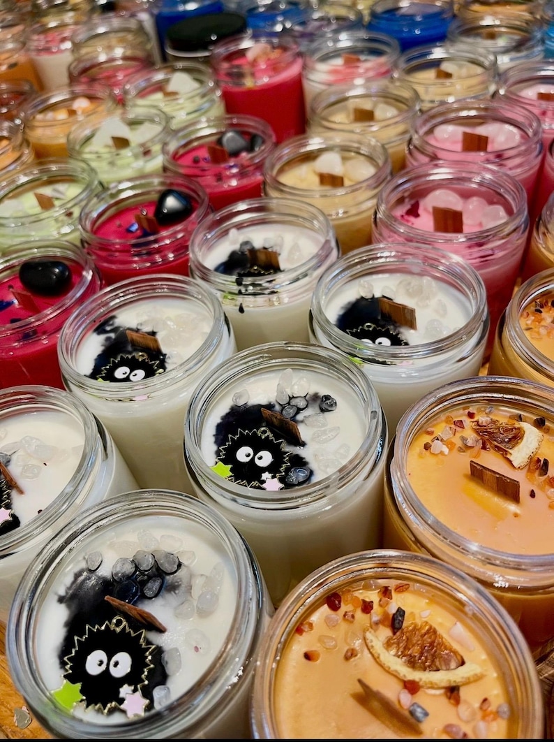 May include: A collection of various scented candles in clear glass jars. The candles feature a range of colors, including pink, red, white, and orange. Some candles have decorative elements like crystals, wood wicks, and cartoon characters.