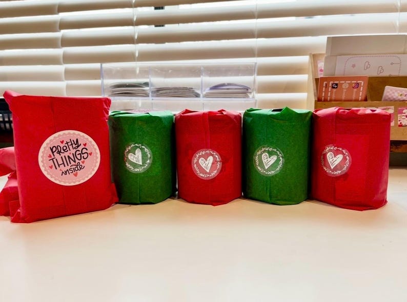 May include: Five wrapped gifts in red and green paper. The gifts are cylindrical and rectangular, with stickers that say "Pretty Things Inside" and heart designs. The gifts are arranged on a white surface.