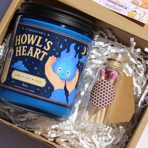 Puede incluir: Un tarro de vela azul con tapa negra, etiquetado "Howl's Heart" con un dise&ntilde;o de llama de dibujos animados. El tarro est&aacute; en una caja de cart&oacute;n con papel triturado, junto a una botella de cristal con cerillas. La vela es de 227g.