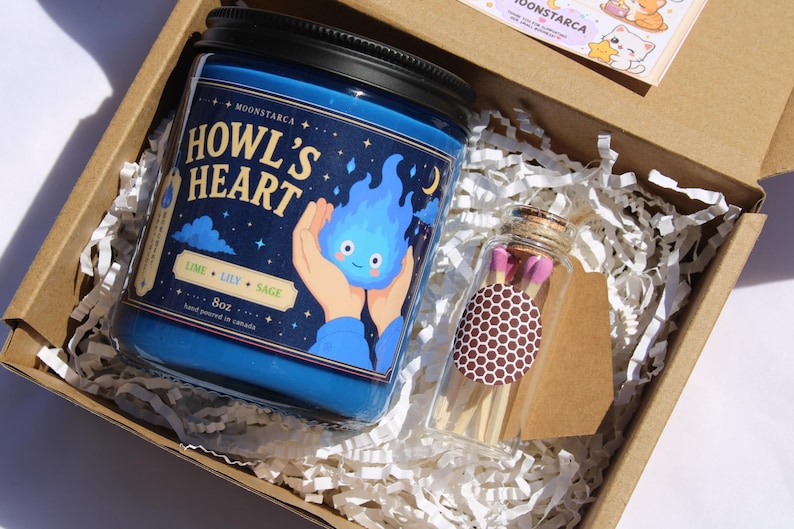 Puede incluir: Un tarro de vela azul con tapa negra, etiquetado "Howl's Heart" con un dise&ntilde;o de llama de dibujos animados. El tarro est&aacute; en una caja de cart&oacute;n con papel triturado, junto a una botella de cristal con cerillas. La vela es de 227g.