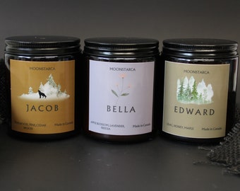 May include: Three glass jar candles with black lids. Each label features a name: Jacob, Bella, and Edward. The Jacob candle has a wolf and forest graphic. The Bella candle has a floral design. The Edward candle has a forest graphic.