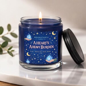 May include: A lit, navy blue scented candle in a glass jar with a black lid. The label reads "A Heart's A Heavy Burden" with the scent notes "White Tea, Rose, Musk". The candle is 8 oz.