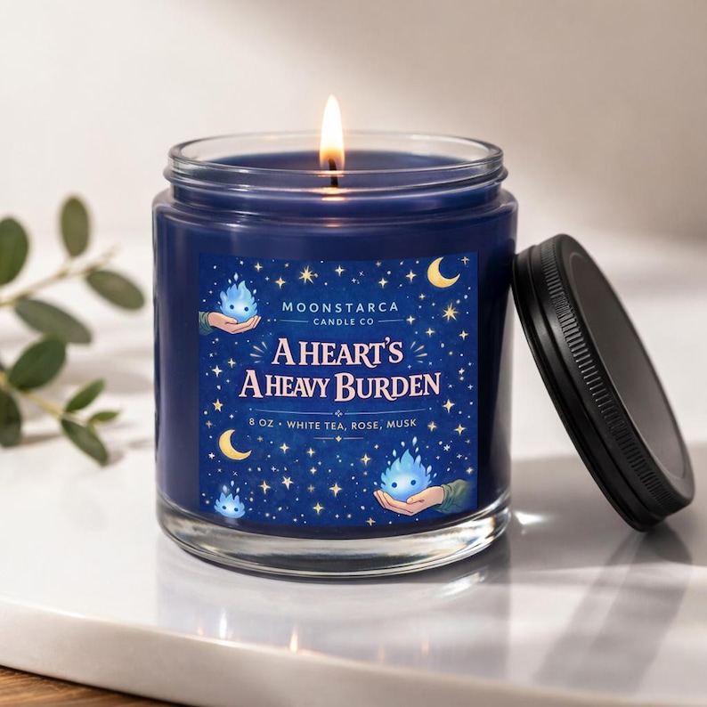 May include: A lit, navy blue scented candle in a glass jar with a black lid. The label reads "A Heart's A Heavy Burden" with the scent notes "White Tea, Rose, Musk". The candle is 8 oz.