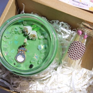 May include: A green candle with decorative elements, including white flowers and a Totoro charm, sits in a glass jar within a cardboard box. A small glass bottle of matches with pink tips and a brown label is also in the box.