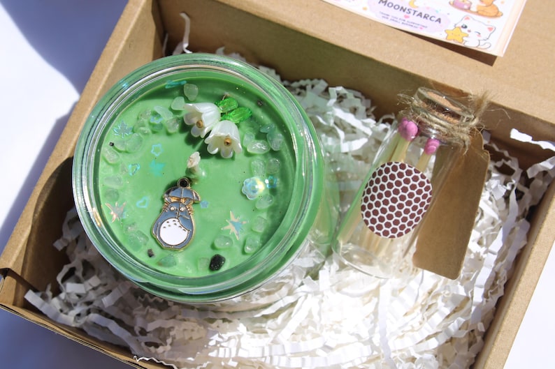 May include: A green candle with decorative elements, including white flowers and a Totoro charm, sits in a glass jar within a cardboard box. A small glass bottle of matches with pink tips and a brown label is also in the box.