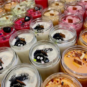 Puede incluir: Una colecci&oacute;n de velas perfumadas en frascos de vidrio transparente. Las velas presentan diferentes colores, incluyendo rosa, rojo y naranja, con elementos decorativos como cristales y mechas de madera. Algunas velas tienen un dise&ntilde;o de personaje de dibujos animados.