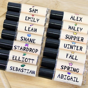 Può includere: Una collezione di balsami per labbra con tappi neri ed etichette con nomi come "Sam", "Emily" e "Alex". Ogni etichetta ha un elemento di design unico. I balsami per labbra sono disposti su una superficie di legno chiaro.