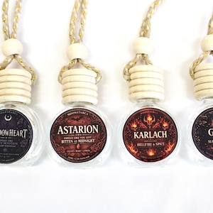 May include: Four clear glass air freshener bottles with wooden tops and decorative labels. The labels feature text like "Shadowheart" and "Astarion" with dark and fiery designs. Each bottle has a gold and beige string for hanging.