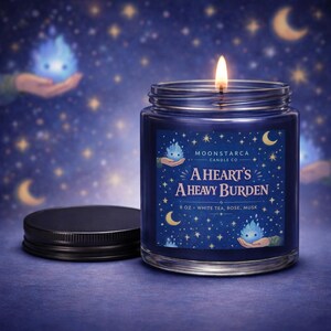 May include: A lit, navy blue scented candle in a glass jar with a black lid. The label reads "A Heart's A Heavy Burden" with the scent notes of white tea, rose, and musk. The background features a starry night sky with crescent moons.