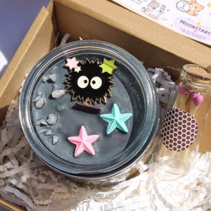 May include: A gift box containing a candle, a small glass bottle, and stickers. The candle is in a clear glass jar with a dark gray wax and decorative starfish and a black character pin. The bottle has a cork stopper.