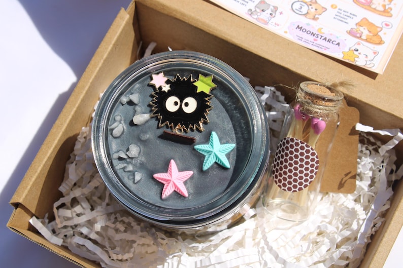 May include: A gift box containing a candle, a small glass bottle, and stickers. The candle is in a clear glass jar with a dark gray wax and decorative starfish and a black character pin. The bottle has a cork stopper.