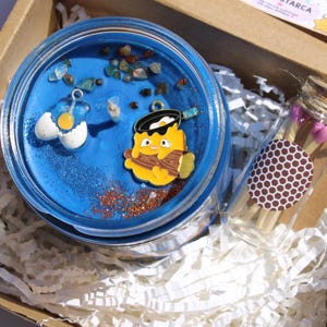 Puede incluir: Una vela azul en un tarro de cristal con abalorios decorativos, purpurina y peque&ntilde;as piedras. La vela est&aacute; empaquetada en una caja de cart&oacute;n con papel triturado y un peque&ntilde;o vial de cristal de cerillas.