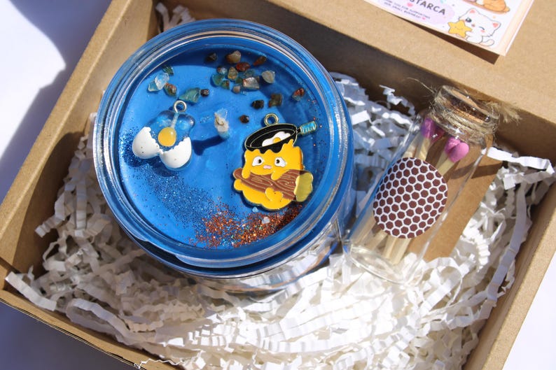 Puede incluir: Una vela azul en un tarro de cristal con abalorios decorativos, purpurina y peque&ntilde;as piedras. La vela est&aacute; empaquetada en una caja de cart&oacute;n con papel triturado y un peque&ntilde;o vial de cristal de cerillas.