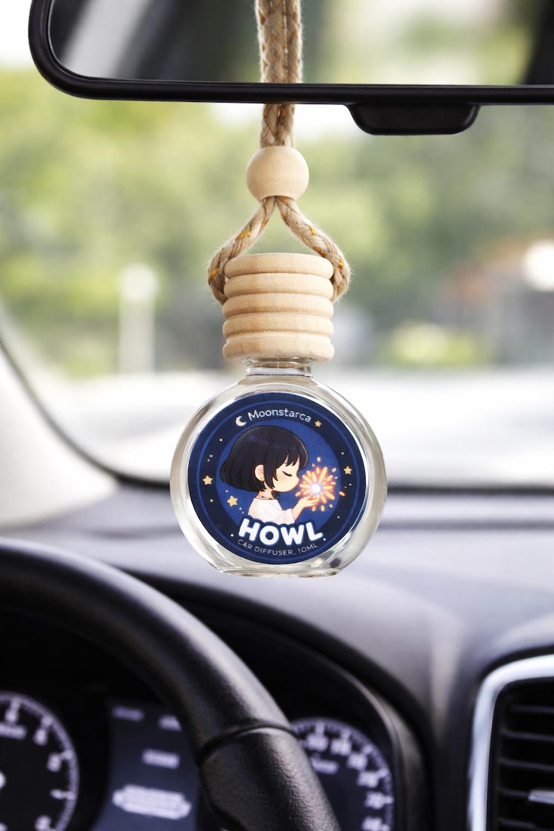 May include: A car air freshener with a round glass bottle and wooden top, hanging from a rearview mirror. The bottle features a blue label with an anime-style illustration and the text "HOWL CAR DIFFUSER".